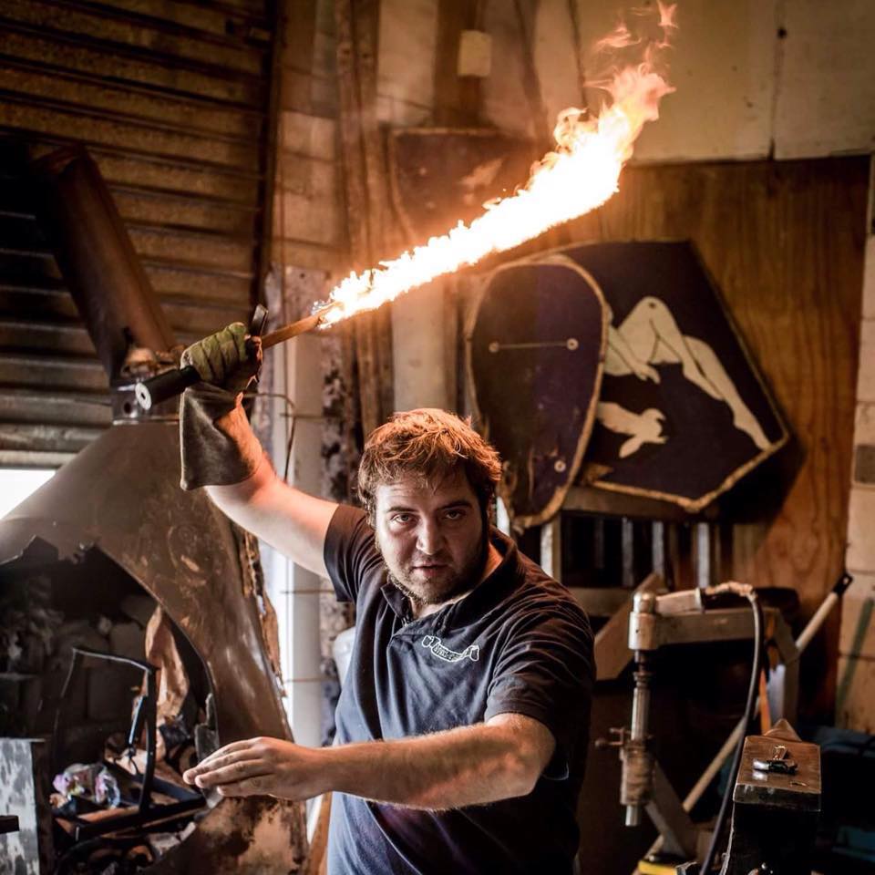 Justin Holding a flaming sword in one of the first workshops, surrounded by tools and medieval armour