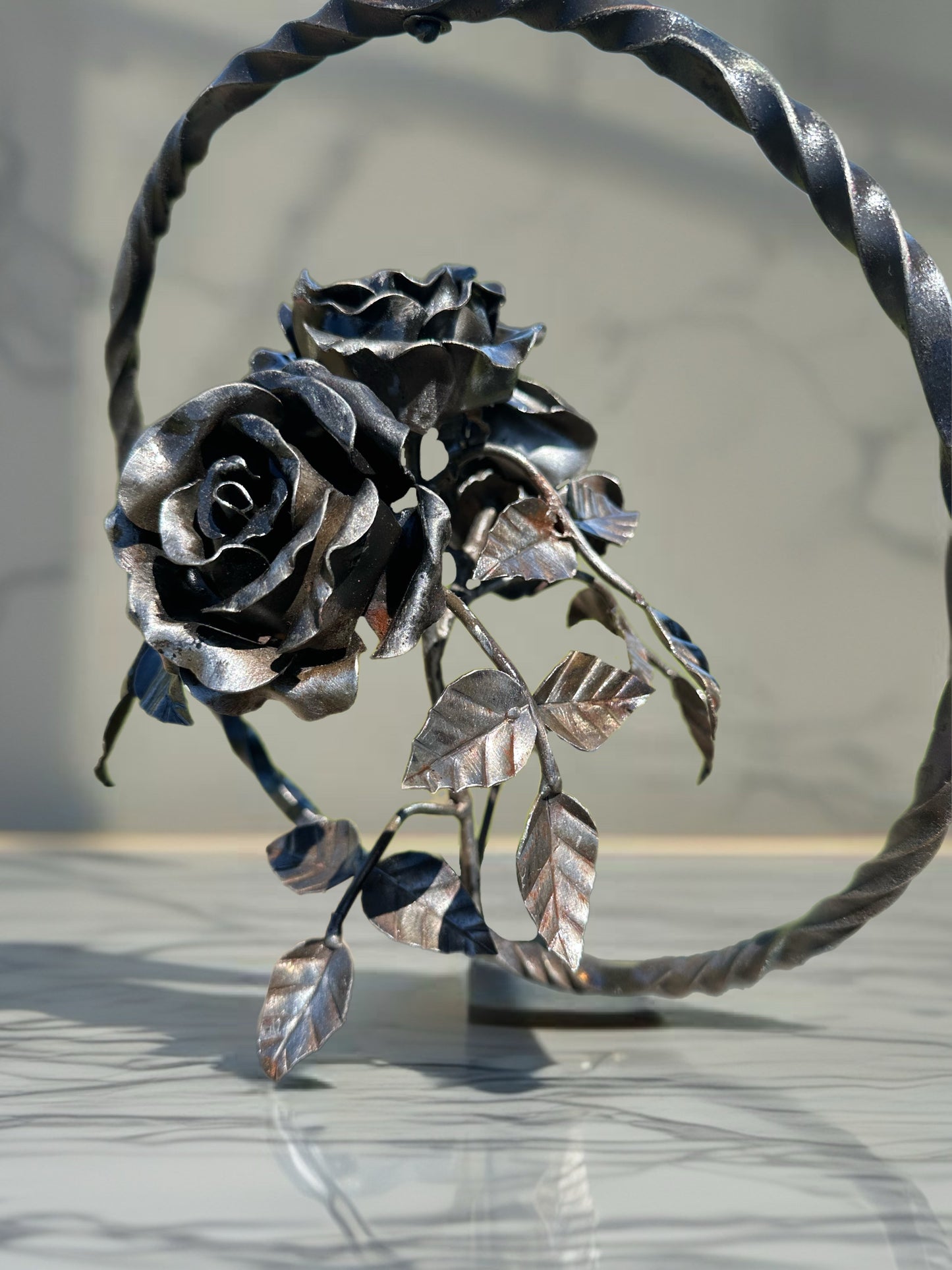 Hand-forged steel garland with three sculpted metal roses arranged on a single stem, displayed inside a circular steel ring against a softly blurred background