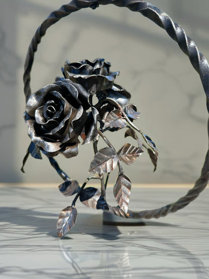 Hand-forged steel garland with three sculpted metal roses arranged on a single stem, displayed inside a circular steel ring against a softly blurred background