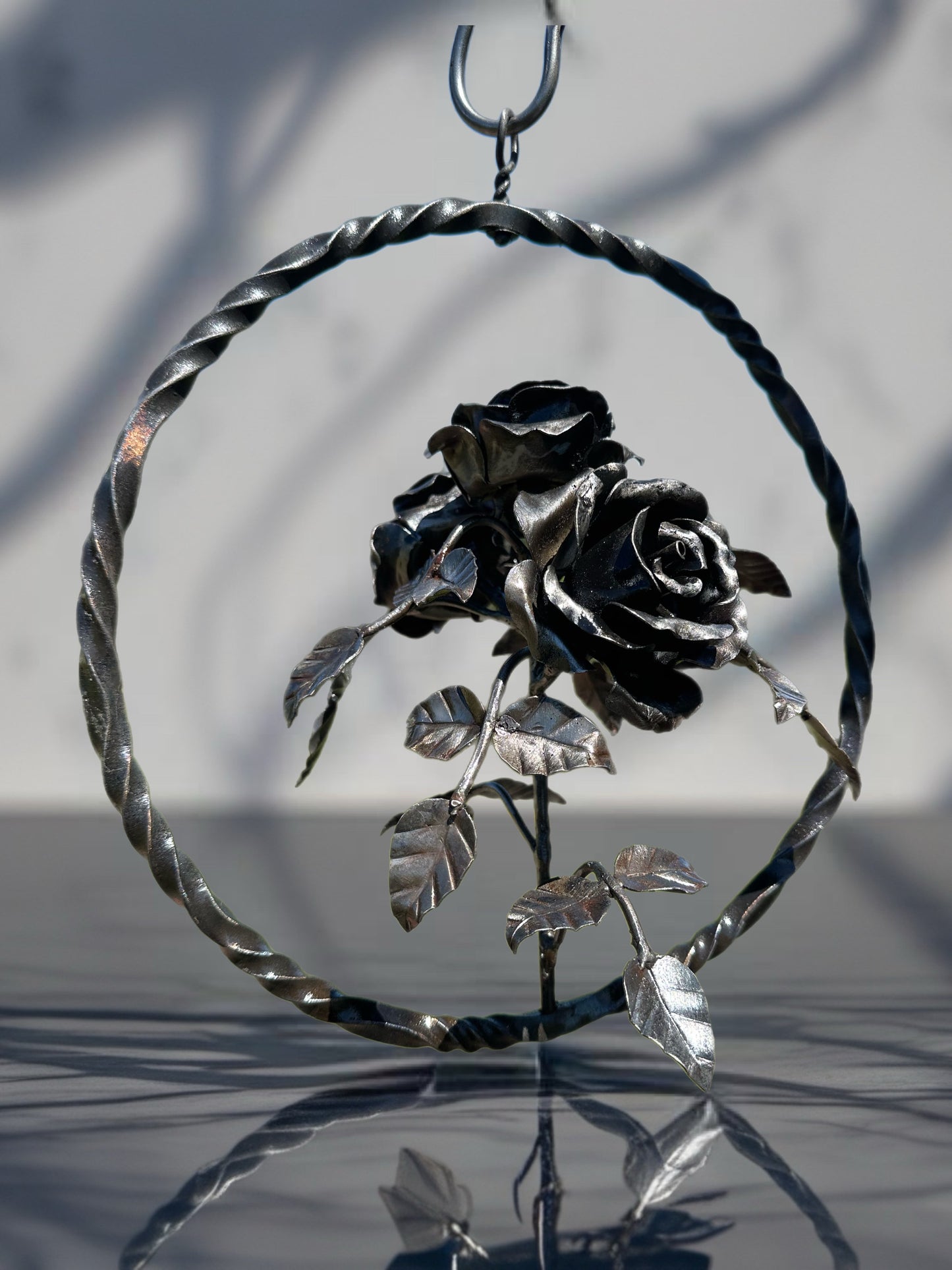 Hand-forged steel garland with three sculpted metal roses arranged on a single stem, displayed inside a circular steel ring against a softly blurred background.